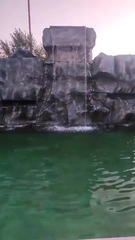 A man-made waterfall cascades into a greenish swimming pool. The waterfall is constructed from rocks, and trees can be seen in the background. The sky is a pale pinkish color, suggesting it might be sunrise or sunset.