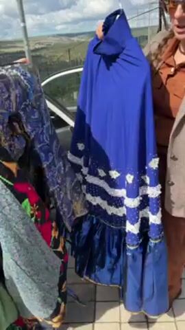 Two women hold up dresses for inspection. One woman on the left wears a green blouse and patterned skirt. She holds a patterned dress with florals. The other woman is on the right and wears a brown jacket. She holds a long, blue dress with white accents and a ruffled hem. There is a black car behind them, and a grassy, hilly landscape in the background. The weather is overcast. The women appear to be discussing the dresses.