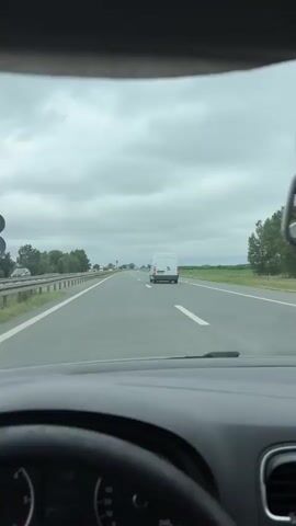 The video shows a first-person view from inside a car driving on a highway. The dashboard and steering wheel are partially visible in the foreground. The road ahead is paved with multiple lanes, separated by white lines. There are other vehicles on the highway, including cars and a white van in the distance. On the left side of the road, there is a guardrail and trees. On the right side, there is a grassy field and some trees. The sky is overcast with heavy clouds. A green highway sign is visible on the right side of the road. The van ahead is moving away from the camera.