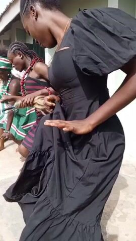 A group of young people, mostly girls, are dancing barefoot outside a building. The girls wear different traditional African clothing, including dresses, skirts, and head wraps in various colors. Some wear beaded necklaces and bracelets. One girl carries a blonde wig, and another wears a baby on her back. A few boys and adults are also present, some dancing and some watching. The building has light green walls and dark green window frames. The ground appears to be concrete. The dancers move to the music, their expressions ranging from focused to joyful.