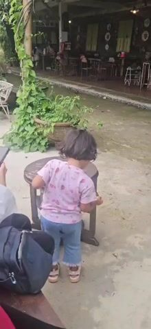 A young child with dark hair, wearing a pink t-shirt with strawberry patterns and blue jeans, is standing near a wooden stool on a concrete floor. The child is holding a yellow object, possibly a leaf, and appears to be interacting with something on the ground. To the left of the child, a black bag is visible on a wooden surface. In the background, there is a covered area with tables and chairs, and lush green plants. The environment suggests an outdoor cafe or restaurant setting.