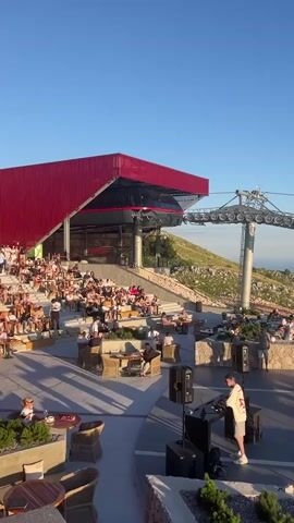 A DJ is seen performing at a mountaintop venue with a large audience seated in an amphitheater-style seating area. A cable car system is visible in the background, extending across the mountain. The venue appears to be a restaurant or lounge, with tables and seating areas set up for patrons to enjoy the view. The sky is clear blue, and the overall atmosphere seems festive.