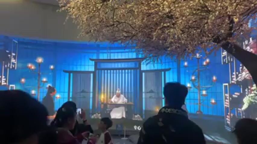 A group of people, some wearing masks, stand in front of a curved blue screen showing a digital projection of a Chinese scholar seated at a desk in front of a traditional building with lit candles on either side. Two men in traditional dress stand on either side of the scholar. Above and to the right is an artificial tree with white blossoms. Some words in a foreign language appear at the bottom of the screen.