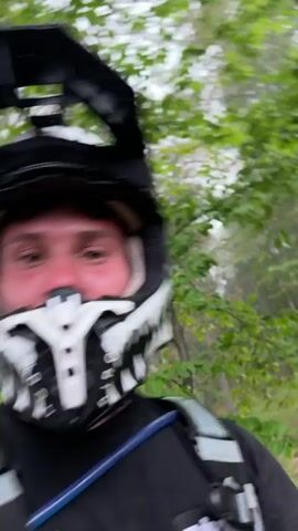 A man in a black and white motocross helmet and black t-shirt with a hydration pack gives a thumbs-up. The camera then pans quickly, showing a person with a neon yellow helmet and then a wide shot of a green, hilly landscape under a misty sky with tree branches in the foreground.