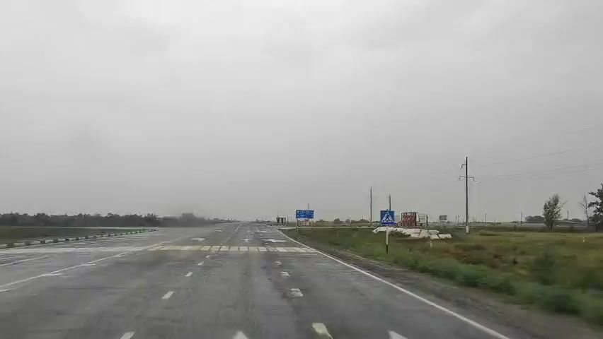 A forward-facing dash cam video captures a highway on a cloudy, rainy day. The road is wet, with visible water on the surface. Lane markings, including arrows and a pedestrian crossing, are visible. On the right side of the road, there are several signs, including directional signs in a foreign language and a pedestrian crossing sign. Electrical poles with wires are also visible. The landscape is flat with some vegetation and trees in the distance. The weather appears to be inclement, with rain possibly affecting visibility.
