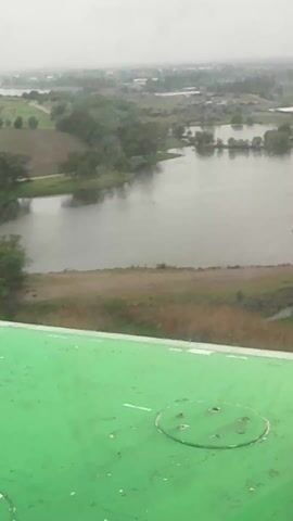 The video presents an aerial view of a landscape featuring a body of water, likely a pond or small lake, surrounded by greenery and some rural infrastructure. The perspective is from a high vantage point, possibly from a building or elevated structure, indicated by the presence of a green, textured surface in the foreground, likely a roof. The weather appears overcast or rainy, giving the scene a muted tone. The body of water is calm, with subtle reflections of the surrounding trees and sky. On the land, there are patches of green grass, some dirt paths, and scattered trees. In the distance, buildings are visible, suggesting a rural or semi-rural setting. The overall impression is one of a serene, damp landscape.