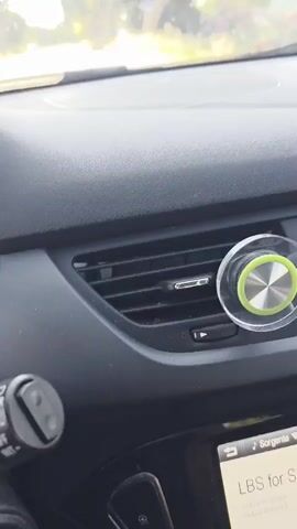 The video shows the interior of a car as it drives along a road. The camera is positioned on the dashboard, facing forward. A green and silver circular object is attached to the air vent. The view outside the car window shows trees, grass, and a bridge structure in the distance. The sky is clear and blue.
