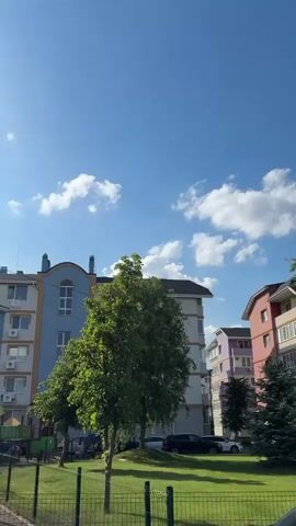 The video displays a sunny daytime view of a modern residential area, likely a housing complex or apartment block. The scene is dominated by several multi-story buildings, characterized by their varied pastel colors including blue, white, pink, and purple, featuring multiple windows and balconies.

Green spaces are prominent, with well-maintained grassy areas and numerous trees; both leafy deciduous trees with bright green foliage and darker coniferous trees are visible. Parked cars can be seen on the paved areas adjacent to the buildings and grass. Briefly, a section of a children's playground with colorful equipment is visible.

The camera performs a slow panning motion, starting by showing the blue and white buildings on the left, then moving across to reveal more of the complex. As it pans, a tall street light post comes into view. The bright blue sky is dotted with scattered white, fluffy clouds. The sun is very bright, especially towards the latter half of the video, creating some lens flare as the camera points more directly towards it. No people or specific activities are observed throughout the video, focusing instead on the architecture, greenery, and sky. The overall impression is one of a calm and pleasant urban environment on a clear, sunny day.