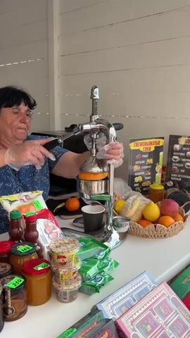 An older woman with dark hair wears a blue floral top and clear gloves. She operates a manual juicer to extract juice from an orange placed in the juicer. The juicer is made of stainless steel. On the table, there is a basket of fruits, including oranges, lemons, and apples, along with jars of honey, packets of sauces, and other items for sale. A menu board displaying "СВЕЖЕВЫЖАТЫЕ СОКИ" (freshly squeezed juices) is visible in the background. The scene is likely a market stall or a small shop.
