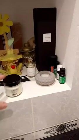 The video shows a white shelf lined with gray-marbled tiles.  The shelf is stocked with decorative and aromatic items.  To the left, a hand places a small white candle with a black lid. The items on the shelf include a black rectangular box with "Aromagraphia" printed on it, several small bottles of oils, a gray candle holder with holes, two small jars labeled "Lilac" and "Peony Blossom," a brown box with the word "ADROP" on it, a yellow wooden flower, and a green frog figurine. The objects on the shelf are clustered together.