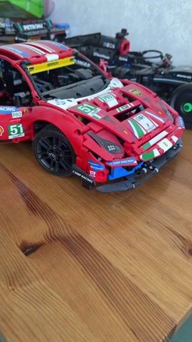 The video shows a Lego model of a Ferrari race car on a wooden surface. The car is primarily red with white, green, and blue accents. It has multiple sponsor logos, including Shell, SMP Racing, and Adler. The driver's side door is open, revealing the interior which is made of black Lego pieces. The tires are black. In the background, there are other Lego models, possibly Formula 1 cars. The wooden surface has a visible wood grain pattern.
