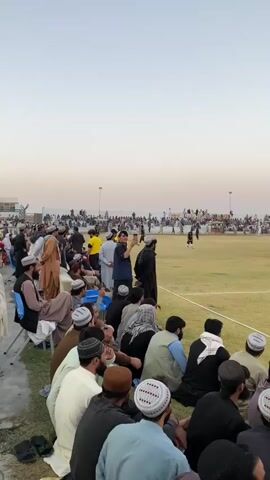 The video shows a wide shot of a soccer game being played on a grassy field. In the foreground, a crowd of spectators sits on the ground, facing the field. They are all men, wearing traditional clothing, including turbans and caps. Some are talking and gesturing, while others watch the game. In the middle ground, players in athletic wear are scattered across the field, some in motion, likely playing the game. The background shows more spectators in bleachers, a city skyline, and a clear sky. The overall atmosphere appears to be that of a local sporting event.
