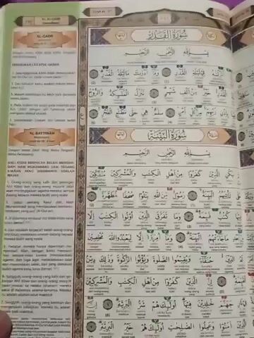 The video pans across an open book, which appears to be the Quran, showing pages with Arabic text and what looks like translations or explanations. The book is laid open, and the pages are densely packed with text and some colored markings or highlights. The lighting suggests an indoor environment. The perspective is from above, looking down at the book. The camera movement is smooth, indicating a deliberate recording rather than a shaky, handheld approach. There are no visible people in the frame, only the book itself. The environment appears to be a table or flat surface.