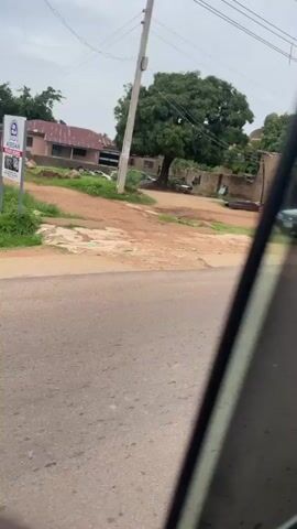 This video captures a view from a moving vehicle on Rukuba Road in Nigeria. The scene unfolds along a dusty road lined with shops and houses. The architecture suggests a semi-urban or rural setting, with many structures featuring corrugated iron roofs and open storefronts displaying various goods, including textiles and what appear to be household items.

Several individuals are visible. In the foreground, a person wearing a red shirt, blue jeans, and a backpack is seen near a parked car, seemingly interacting with someone inside or preparing to move. Further down the road, other people are seen around the shops, though their activities are less distinct due to the distance and movement of the vehicle. Children are also present, with two boys in one shop engaging in an activity.

The time of day appears to be overcast, with diffused natural light suggesting either early morning or late afternoon, or a cloudy day. The weather seems mild, and the presence of greenery indicates a period outside of severe drought. The overall impression is of a typical day in a Nigerian community, with daily commerce and pedestrian activity.

Notable details include the parked cars, bicycles, and the variety of merchandise displayed outside the shops. One shop has "BROWN'S FEED" visible on a sign. The road itself is unpaved in areas, typical of many roads in Nigeria, and shows signs of recent activity. The presence of a prominent rocky outcrop in the background adds a distinctive geographical feature to the landscape. The video is shot from a slightly elevated perspective within a vehicle, implying the viewer is a passenger.
