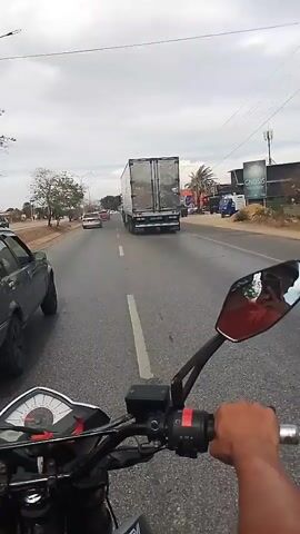 A motorcycle is driving on a wet, asphalt road. The camera is mounted on the handlebars of the motorcycle, providing a first-person perspective. To the left of the motorcycle is a dark gray car, and to the right is a large, white semi-truck. Other cars are visible in the distance. The sky is overcast. Palm trees are visible in the distance to the right. The rider's right hand is on the throttle.