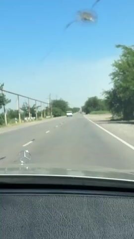This video shows a first-person perspective from inside a car, traveling down a paved road. The dashboard and the Mercedes-Benz emblem on the hood are visible in the foreground. The road stretches ahead, with a white line dividing lanes and a solid white line marking the shoulder.

On the left side of the road, there are power lines supported by poles and a metal fence. Beyond the fence, there are trees and some greenery. On the right side of the road, the terrain appears drier with sparse vegetation and some trees further in the distance.

Several vehicles are visible on the road ahead. A white van is seen in the distance on the left lane, and other cars are further down the road. The sky is clear and blue, indicating a sunny day. The overall scene suggests a rural or suburban setting during daylight hours. There is a notable crack or damage on the windshield in the upper right portion of the frame.

There is no visible text in the video. The activity is a car journey on a road. The weather appears to be warm and sunny. The location cues suggest a road in Armenia, possibly Surenavan, Armenia as indicated by the metadata.