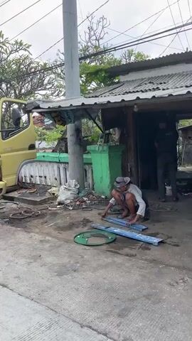 A man wearing a hat and a light-colored shirt crouches on the ground, working with metal pipes. He is positioned in front of a small, open-fronted building with a corrugated metal roof. Behind him, a yellow truck is partially visible. To the right of the building, a display table covered with a blue and white patterned cloth is set up, featuring various items for sale, including what appears to be food. The ground is a mix of dirt and asphalt. The overall environment suggests a roadside stall or workshop.