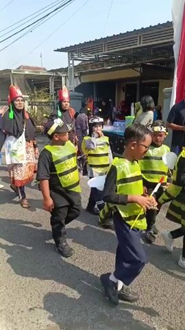 A group of people, primarily women and children, are walking in a procession on a paved street. The women are wearing black hijabs and elaborate, colorful dresses, some with patterned fabrics. They are also wearing tall, conical red and gold hats, and some have large, novelty sunglasses. The children are dressed in black shirts and vests with yellow and black stripes, resembling bees. Some of the children are wearing yellow caps. The procession appears to be taking place outdoors during the day, with houses and buildings visible in the background. The sky is clear and blue, suggesting sunny weather. The people are walking in the same direction, and some are waving. The overall scene suggests a festive event or parade.