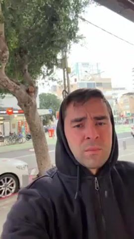 The video shows a man in a black hooded sweatshirt standing outside. He is looking directly at the camera, his face showing a neutral expression. A tree is behind him to the left. A sidewalk runs in front of him, with a white car parked at the curb. A bicycle rack is visible with several bicycles in it. Across the street, there are buildings and shops. Pedestrians can be seen walking. The sky is overcast.