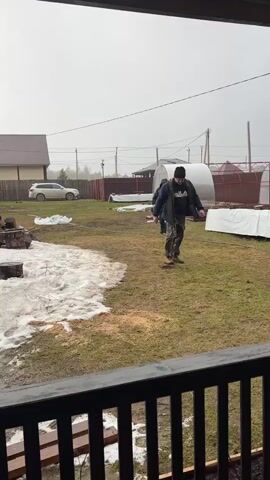 Two men are walking across a grassy, muddy yard on a grey, overcast day. One is wearing a black knit cap, a dark blue Fila sweatshirt, a grey vest, and camouflage pants. The second man is wearing a hooded camouflage jacket and dark pants. Behind them are two small greenhouse structures, a brown fence, and a house in the background. There are patches of melting snow on the ground. The video is shot from a porch with a black railing. The men are walking towards the camera.