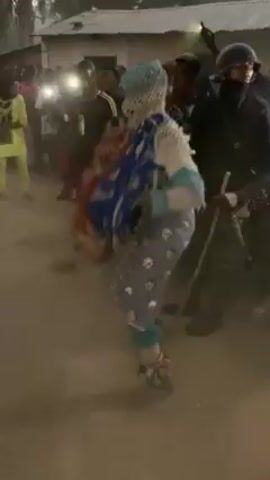 The video shows a masked figure performing a dance in a dusty outdoor setting. The figure is dressed in an elaborate, multicolored costume with a blue patterned backpack. The figure is surrounded by a crowd of onlookers, some of whom are holding up cell phones to record the performance. A police officer stands nearby with a baton. In the background, there are buildings and trees. The overall atmosphere suggests a festive or cultural event.