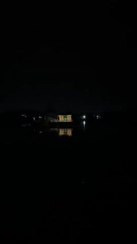 A shot of a houseboat at night. The houseboat is illuminated by interior lights, casting a warm glow on the water. The water is calm and reflects the houseboat and its lights. The sky is dark and clear, with a few distant lights visible.