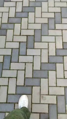 The video shows a first-person perspective of someone walking on a patterned walkway. The walkway is made of interlocking rectangular pavers, alternating in light gray and dark gray colors, arranged in a herringbone pattern. The person is wearing white sneakers and green camouflage pants. The camera angle is angled downwards, focusing on the feet and the ground. The footage is shaky, indicating movement.
