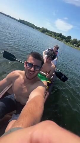 Three young men are in a canoe on a lake. The man in the front is holding the camera, creating a selfie-style view. He is shirtless, wearing sunglasses and a gold chain, and has a short beard. The second man is also shirtless and wearing green shorts. The third man is wearing a white shirt and what appears to be a life vest. Each man has an oar. The lake is wide and calm with visible shore in the distance. The sky is blue with scattered clouds.