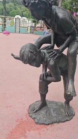 A bronze sculpture of two children is the central focus of the video, positioned on a textured red ground. One child is bent over, with the other child riding piggyback. Their figures are wet, with water droplets visible on the sculpture. In the background, a playground with swings and other equipment can be seen. Several people, including children and adults, are present in the playground area, some interacting with the equipment. The environment appears to be outdoors, possibly a park or recreational area, with trees and a railing visible in the distance.