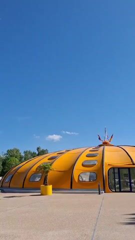This is a short video clip filmed on a bright, sunny day. The camera pans across a bright orange, dome-shaped building with multiple oval windows. A small palm tree in a yellow pot sits near the entrance. The building appears to be part of a larger complex, as indicated by the surrounding paved area and some trees and greenery in the background. Further in the distance, a restaurant with outdoor seating is visible, along with bicycles parked nearby. The sky is clear blue with a few wispy clouds. The overall atmosphere is relaxed and pleasant, suggesting a warm day in a recreational or tourist location. The time of day appears to be midday, given the bright sunlight and clear skies. There are no discernible text elements visible in the video.