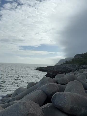 This is a video clip of a coastal scene in Sesimbra, Portugal. The camera pans across a view of the ocean from a rocky shoreline. In the foreground, large, concrete tetrapods are piled up, forming a breakwater. The sea is a deep blue, with gentle waves lapping against the rocks. The sky is partly cloudy, with patches of blue visible between the white clouds. In the distance, cliffs and trees can be seen along the coastline. The lighting suggests it is daytime, likely mid-morning or afternoon, with soft sunlight filtering through the clouds. There is no visible text in the video. The overall mood is peaceful and serene, showcasing the natural beauty of the Portuguese coast.