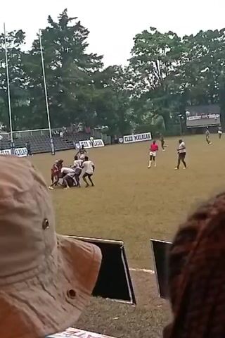 The video shows a rugby game being played on a grassy field. Several players are in a scrum, while others are positioned around them. Some spectators are visible in the bleachers in the background, as are trees. The video is recorded from a vantage point behind an individual wearing a tan bucket hat and another individual with braided hair. Signage displaying "CLEO MALALA" is visible along the bleachers. The lighting is overcast, suggesting it could be a cloudy day.