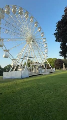 The video shows a large, white Ferris wheel in a park setting. The wheel is stationary, with empty passenger cars hanging from it. The base of the wheel is surrounded by white barriers with logos. The ground is covered in green grass, and there are trees in the background. The sky is blue. There are a few people working on the wheel, but they are not the main focus of the shot.