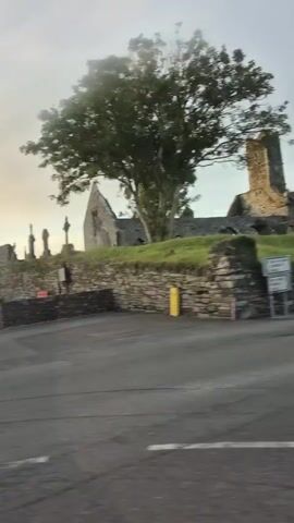 The video shows a view from a moving vehicle looking towards a rural village. The time appears to be early morning or late evening due to the low light and soft sky. In the foreground, there is an asphalt road. Beyond the road, a low stone wall separates it from a grassy area. On the grassy area, there are some buildings and a large tree.

In the first few seconds, the camera pans across a scene that includes a white van parked near the stone wall and some ruins in the background. There is a tall wooden pole with electrical wires and a street light visible.

As the video progresses, the view shifts to show several houses with typical rural architecture, including pitched roofs and chimneys. Some of the houses are painted in pastel colors like yellow and green. A dark green electrical box is situated near the road. A van is also visible parked on the street.

The camera continues to move, and the focus shifts to a yellow building with several windows and solar panels on its roof. This building appears to be a school or a community center. The windows show some contents inside, and the building is bordered by another stone wall.

The video concludes by showing more of the yellow building with the solar panels, still viewed from the moving vehicle. The sky remains overcast with soft light. There are no people clearly visible in the footage, and the actions are limited to the movement of the vehicle and the panning of the camera. The environment is that of a quiet, rural settlement.