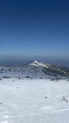 The video shows a snowy mountain landscape under a clear blue sky. Patches of snow cover the ground, with some sparse vegetation peeking through. In the distance, another mountain peak is visible, partially covered in snow and trees. The camera pans slightly, revealing more of the snow-covered terrain and a few small, scattered trees. The scene appears calm and serene.