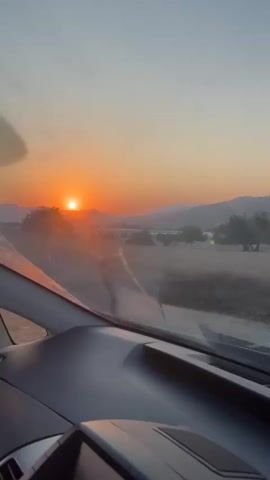 The video shows a view from inside a car, looking out the windshield. The sun is setting, casting a warm orange glow over the landscape. The car is driving on a rural road, with dry grass, trees, and hills visible on either side. Power lines and poles run alongside the road. The dashboard of the car is visible in the foreground. The video appears to be shot in the late afternoon or early evening.