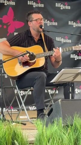 A man with glasses and a black striped shirt sits on a folding chair playing an acoustic guitar and singing into a microphone. He is wearing black pants and black shoes. The guitar is a light brown acoustic guitar with a pickguard. He is wearing a strap over his right shoulder. A music stand is positioned to his right. Behind him is a black banner with pink butterfly graphics and the text "BAKGARDEN" repeated multiple times. In front of him, there is a patch of green grass. The lighting appears to be natural light, suggesting an outdoor setting. The man is focused on his performance, with his eyes closed at times and his mouth open as he sings.