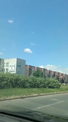 This media file captures a view from a moving vehicle on a sunny day in Ciechanów, Poland. The scene is dominated by a row of identical, light-colored apartment buildings stretching along the right side of the road. These buildings have multiple floors with evenly spaced windows and balconies. A distinct white building with windows and what appears to be a sign is visible on the left.

In the foreground, the road surface is visible, suggesting the perspective of someone driving or riding in a car. The middle ground features a green grassy area with bushes and trees, separating the road from the apartment buildings. Several cars are visible, including a blue SUV, a silver hatchback, and a white van, parked or driving along the road and in what appears to be a parking lot. One car, a dark SUV, is driving ahead of the vehicle from which the recording is made.

The sky is a clear, bright blue with a few scattered white clouds, indicating fair weather and likely daytime. Road signs are present, including what looks like a pedestrian crossing sign and other traffic signals. The presence of apartment buildings, a paved road, and streetlights suggests an urban or suburban residential area. The overall mood is calm and ordinary, depicting a typical day in this Polish location. There is no discernible text on the majority of the visible objects, except for a sign on the white building on the left which appears to have Polish text, and a license plate on the blue SUV which also appears to be Polish.