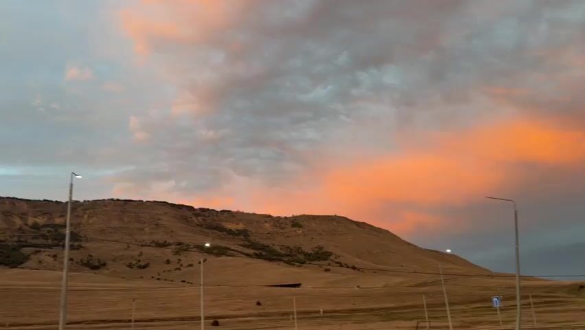 The video shows a wide shot of a barren, hilly landscape with a road and streetlights in the foreground. The sky above is filled with dramatic clouds, illuminated by the warm hues of an impending sunset or sunrise. The clouds are a mix of orange, pink, and gray, with patches of blue sky visible. The hills are covered in dry grass and sparse vegetation. The overall impression is one of a vast, open, and somewhat desolate natural environment at dawn or dusk.