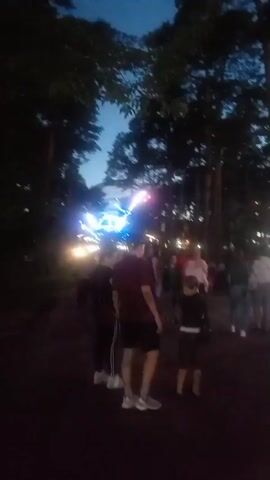The video shows a nighttime outdoor scene with a bright, pulsating blue and white light source in the center of the frame, surrounded by silhouetted trees. Several people are visible in the foreground and background, appearing to observe the light. Some individuals are standing close to the light, while others are further back. The sky above is dark, suggesting it is late evening or night. The light source is large and appears to be a form of illumination or display, possibly at an event.