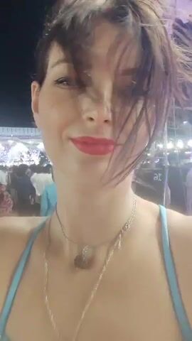 A woman with brown hair, parted on the side and somewhat windswept, takes a selfie video. She is wearing red lipstick and a light blue-strapped top, accessorized with two necklaces. She smiles pleasantly at the camera, with a small glitter accent near her left eye.

The background reveals a bustling outdoor gathering at night in Askihala, India, illuminated by numerous artificial lights. A large crowd of people, dressed in various outfits including some traditional Indian attire, fills the area behind her. The setting is elaborately decorated with white structures, possibly tents or stages, adorned with string lights and chandeliers. The ground is covered with a red patterned carpet or mat. Loud music, appearing to be Indian, plays throughout the venue. A speaker box with the partial text "at" is visible to the left of the woman. The overall atmosphere suggests a festive event, likely a wedding or large celebration.
