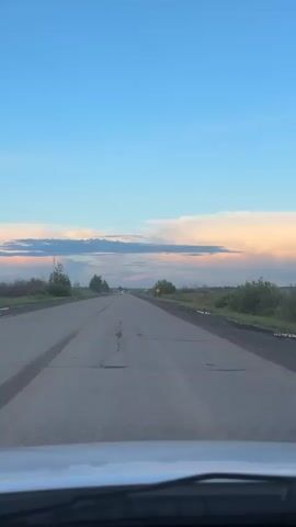 The video shows a view from the interior of a vehicle driving on an empty, paved road. The sky is a clear blue with wispy clouds tinged with pink and orange, suggesting it is either sunrise or sunset. The road stretches ahead into the distance, with some cracks visible on the asphalt. On either side of the road are fields with sparse vegetation and scattered trees. There are two yellow circular signs visible on the right side of the road, one further in the distance than the other. The perspective suggests the video is recorded from the driver's seat.
