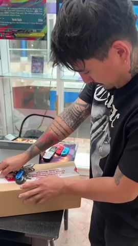 A person with tattoos on their left arm and shoulder is unboxing a cardboard package on a dark table. They are using a small blue tool with keys attached to it to cut the packing tape. There are other objects on the table, including a laptop, a cellphone, and two game controller attachments. The background appears to be a store with glass display cases filled with merchandise. The lighting is bright, and the scene is in focus.