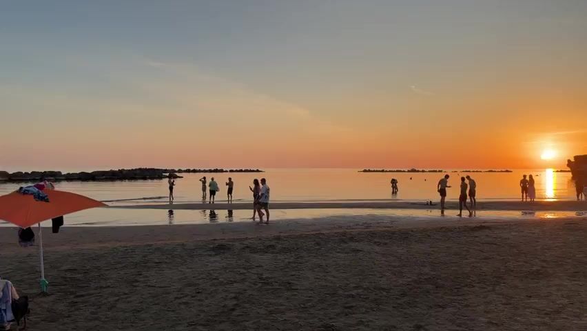 The video shows a beach scene at sunset. The sky is a vibrant orange and pink gradient, with the sun low on the horizon and reflecting on the calm water. Silhouetted figures of people are visible on the wet sand near the water's edge, some walking and some standing. In the foreground, there is a beach umbrella with some items hanging from it. The sand is dark and textured. Further out in the water, more people are scattered, some appearing to be playing or wading. The background shows a rocky coastline on the right and breakwaters in the distance. The overall atmosphere is serene and tranquil.