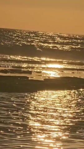 The video shows a close-up view of ocean waves crashing against rocks during sunset. The sun's golden rays reflect off the water, creating a shimmering effect. The sky is a soft orange hue, indicative of the late hour. The rocks appear dark and wet, contrasting with the bright water. The waves are gentle, with a soft lapping sound audible. No people or animals are visible in the frame.