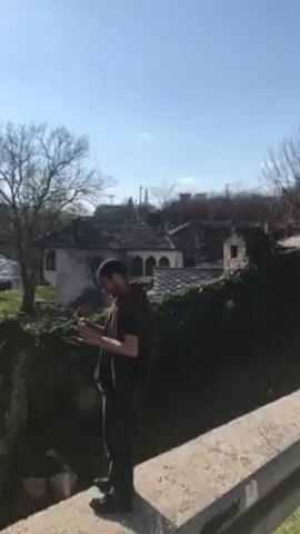 The video shows a man standing on a concrete ledge in front of stone buildings with a view of a river and a mountain in the distance. The man is wearing a dark t-shirt, dark pants, and a jacket over his shoulders. He is holding a small object, possibly a phone, in his hands and looking at it. The stone buildings have dark tile roofs and are surrounded by greenery. The river flows through a green meadow with several trees in bloom. A mountain is visible in the background under a bright, clear sky.