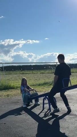 The video shows two people on a seesaw. On the left is a young girl with long blonde hair wearing a blue t-shirt and jeans. She is gripping the handles of the seesaw and smiling. On the right is an adult man with short brown hair wearing a black t-shirt and black pants. He is also gripping the handles. The seesaw is in a playground with a black surface. In the background, there is a green grassy field with a white soccer goal and trees beyond. The sky is blue with some white clouds, and the sun is shining. The people are moving up and down on the seesaw.