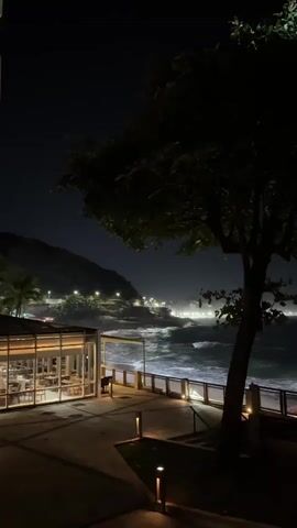 The video captures a night scene from a slightly elevated perspective overlooking a coastal area. In the foreground, a tree with dark foliage is prominent on the right. To the left of the tree, there's a modern building with large glass windows, showcasing an interior with tables and chairs, possibly a restaurant or cafe, illuminated by warm lights. Further left, a tall, white multi-story building with balconies is visible. In the midground, beyond the building with glass windows, a dark concrete or paved walkway extends along the coast, with several illuminated ground lights. A low fence or railing separates the walkway from the ocean. The ocean is active with white-capped waves crashing against the shore. In the background, a dark landmass or hill is visible, dotted with numerous bright streetlights or building lights, indicating a populated area along the coastline. The sky is dark, suggesting night. The overall impression is a vibrant yet serene nocturnal coastal landscape.