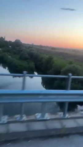 The video shows a view from a bridge at sunset. A river runs below the bridge, with trees and fields on either side. The sky is a gradient of blue and orange, with a few clouds. The camera is moving, suggesting the video was taken from a vehicle traveling across the bridge.