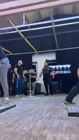 The scene takes place in a large room with a high ceiling and dark curtains around the perimeter. In the center of the room, there's a small round table with two metal bowls on it. Three men are visible: one with a shaved head and wearing blue sweatpants, standing next to the table, one sitting on a black couch with a gray shirt and shorts, and a tall man with a long beard and black tank top standing opposite the man with blue sweatpants. A fourth person stands behind the man in sweatpants. The man with the long beard is speaking to the man with blue sweatpants. There is a TV screen with multiple feeds on the right side, and workout equipment can be seen. The floors are a light color. A light is placed overhead.